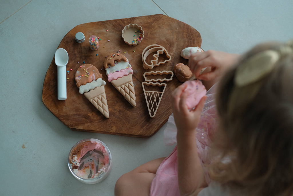 A child enjoys sensory play by decorating cookies shaped like ice cream cones on a wooden board, using icing and sprinkles. Next to them on a light surface are a spoon, the Ice Cream Cutter Set - Eco Playdough Tool, other eco-friendly playdough accessories, and a bowl of pink and white icing.