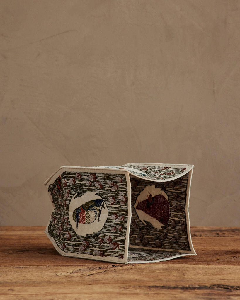 The Coral & Tusk Birdhouse Tissue Box Cover, a small rectangular embroidered cover with playful animal designs, rests on its side atop a wooden surface against a beige textured wall.
