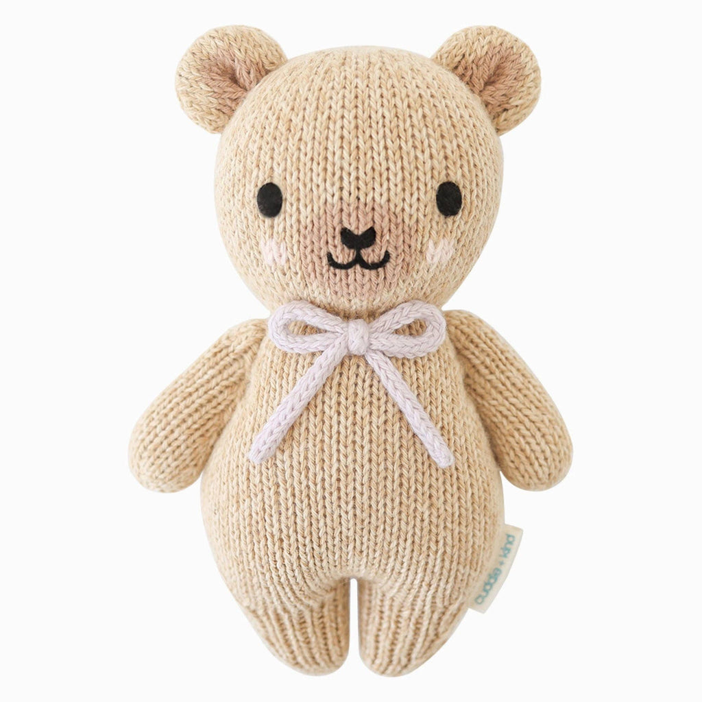 A beige hand-knit Cuddle + Kind Baby Honey Bear with black eyes, a black nose, light pink cheeks, and a lavender bow around its neck sits upright against a white background.
