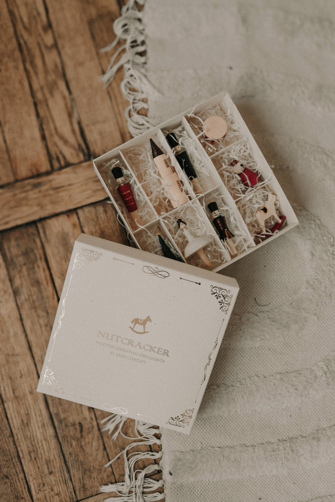 An open Sabo Concept Christmas Ornaments box rests on a wooden floor, displaying hand-painted ornaments and a nutcracker ornament set nestled in shredded paper, with part of a fringed white rug nearby.