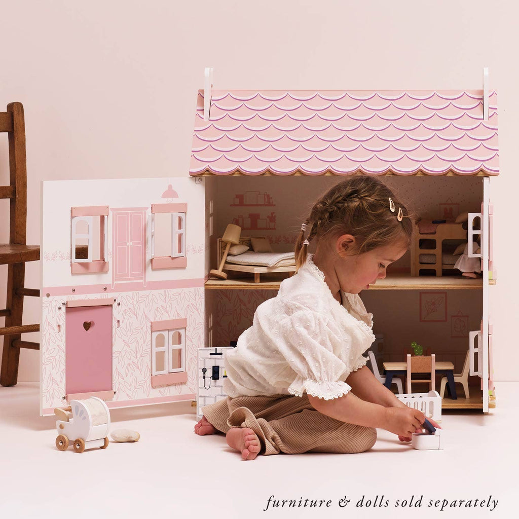 A young child plays on the floor with Sophie’s Wooden Dolls House, crafted from FSC-certified wood and open to show miniature furniture inside. Text at the bottom reads, "furniture & dolls sold separately.
