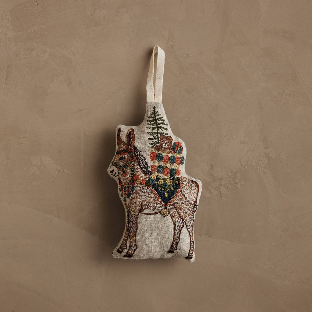 Coral & Tusk Christmas Donkey ornament embroidered on natural linen, featuring a small pine tree and colorful wrapped gifts strapped to its back.