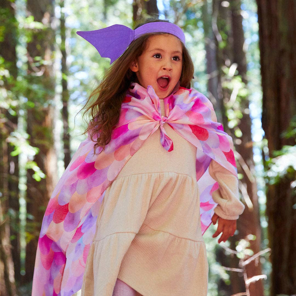 A young child in a flowing dress and purple headband joyfully runs through a sunlit forest, wearing Sarah's Silks Magical Creatures Playsilk - Pink Unicorn, embracing open-ended play.