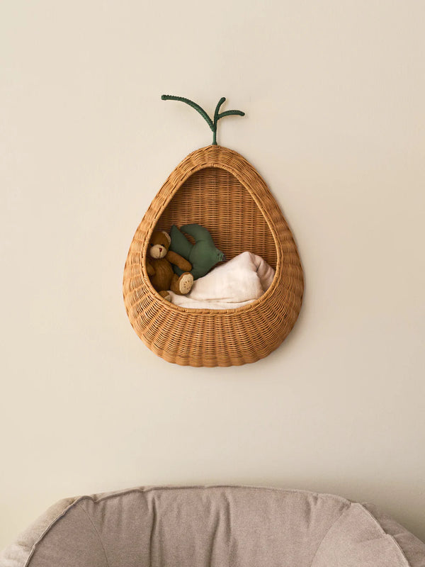 The Ferm Living Pear Braided Wall Pocket - Natural holds a green soft toy, brown teddy bear, and white blanket, hanging on a neutral wall above beige cushioned furniture to add charming rattan storage to your space.