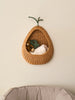 The Ferm Living Pear Braided Wall Pocket - Natural holds a green soft toy, brown teddy bear, and white blanket, hanging on a neutral wall above beige cushioned furniture to add charming rattan storage to your space.