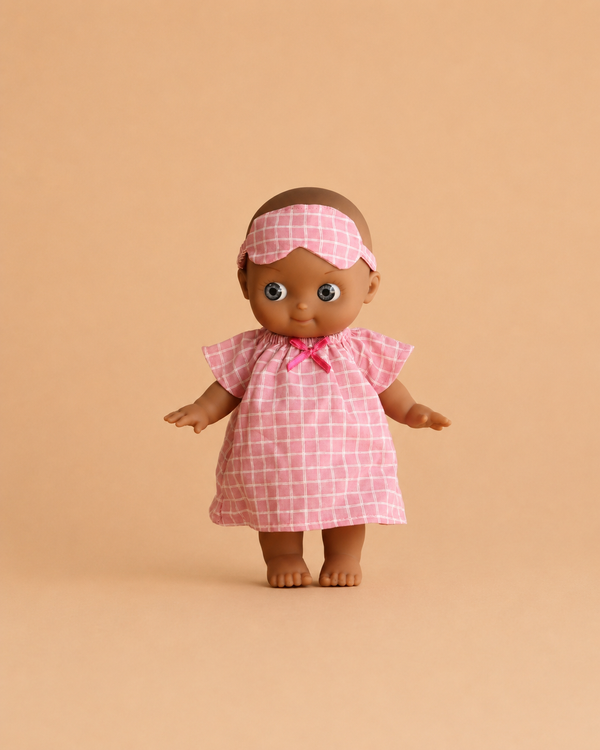The Minikane Mini Pancha Doll - Maia with brown skin is dressed in a pink and white checkered nightgown with a matching sleep mask, set against a beige background—perfect for inspiring imaginative play.