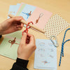A person uses the Knot Game to practice tying complex knots with a red rope and wooden dowel, following illustrated cards. On the table are extra ropes, a metal carabiner, and more instructional cards from the Knot Game set.