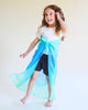A young child with long curly hair smiles barefoot, wearing a white shirt, dark shorts, and a Sarah's Silks Enchanted Playsilk - Sea tied as a cape—perfect for open-ended play and imaginative adventures.