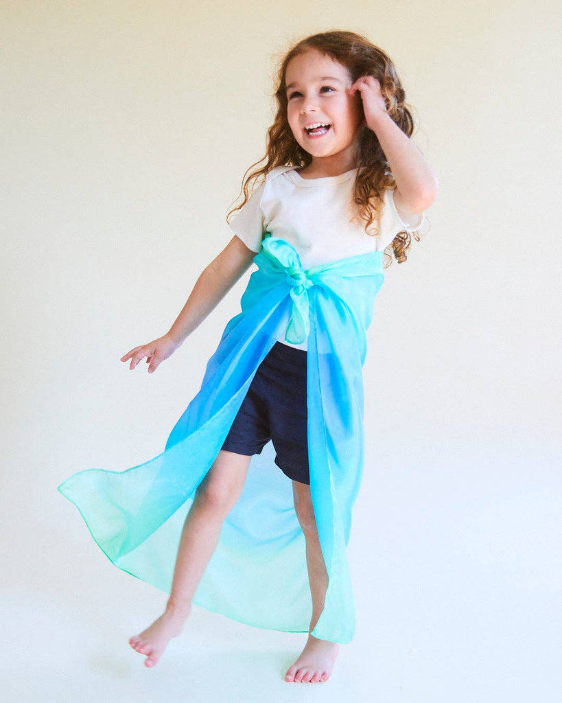 A young child with long curly hair smiles barefoot, wearing a white shirt, dark shorts, and a Sarah's Silks Enchanted Playsilk - Sea tied as a cape—perfect for open-ended play and imaginative adventures.