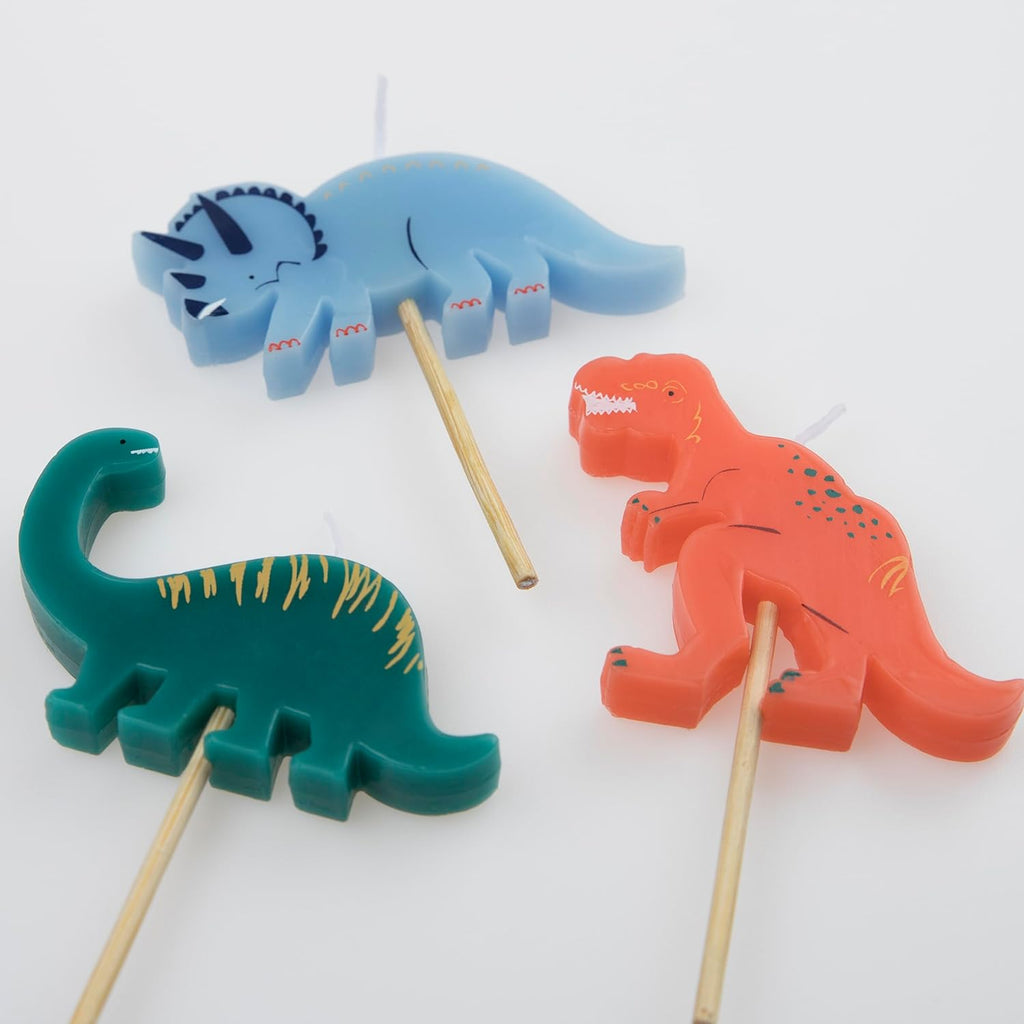 Three colorful Meri Meri Dinosaur Candles in blue, green, and red on sticks, arranged on a white background.