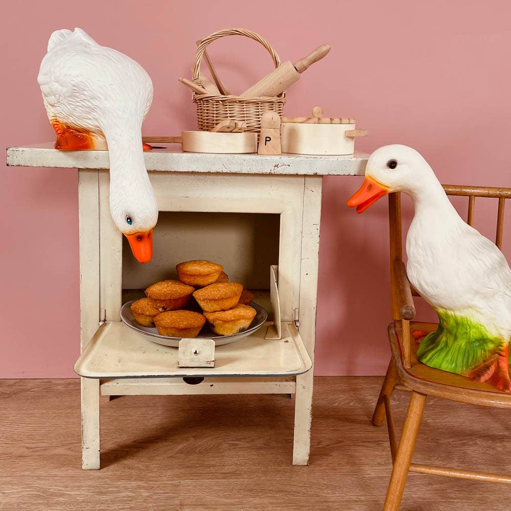 Two Duck Lamps, ideal for children's room decor, explore a small vintage oven—one on a chair, the other peering at muffins inside. Baking tools and baskets sit atop the oven, all set against a charming pink wall.