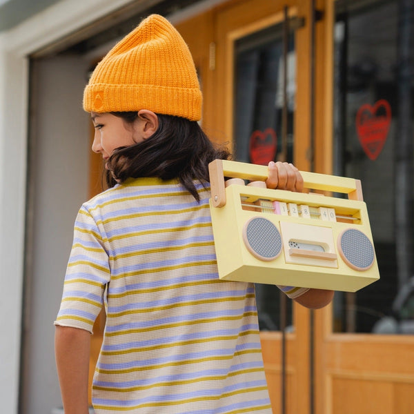 A young girl wearing a striped shirt and orange beanie carries a Retro Wooden Tape Recorder with a handle, walking by a door with heart decorations. She plays her favorite songs on an old school boombox