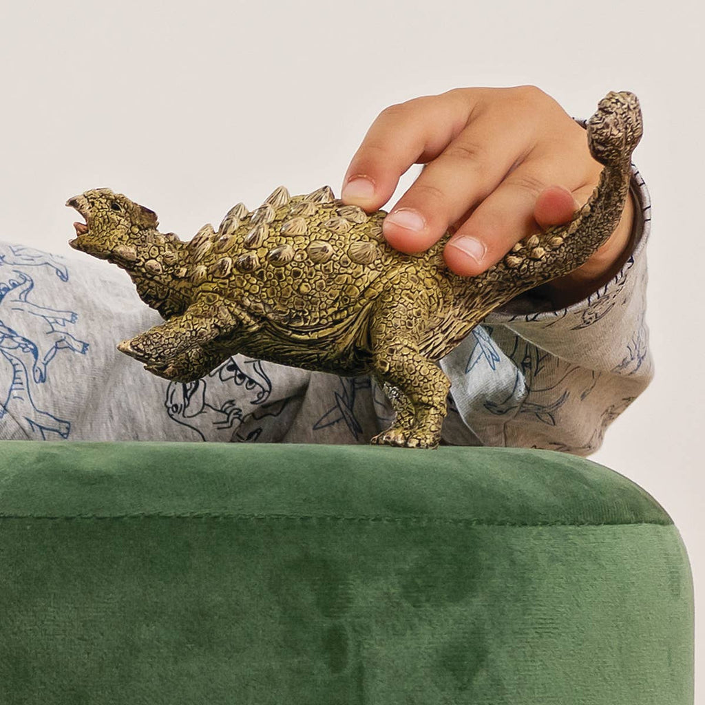 A child’s hand holds the Schleich Ankylosaurus Realistic Dinosaur Toy on a green, plush surface. Wearing a light shirt with blue dinosaur prints, the child adds extra fun and imagination to this educational playtime adventure.