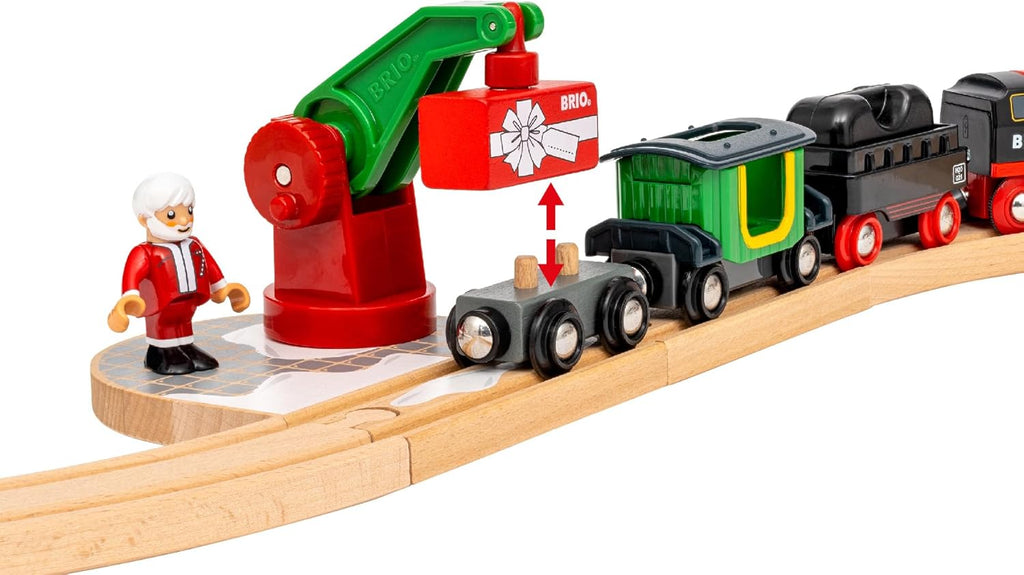 The BRIO World Christmas Steaming Train Set features a red and green crane lifting a gift onto a carriage, with a Santa figure standing on the track beside the black and red steaming train engine and carriages.