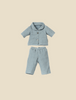 Introducing the Maileg Extra Clothing: Pyjama for Teddy Dad, a delightful doll ensemble that features a light blue short-sleeve button-up shirt paired with matching pants, all adorned with a subtle gray striped pattern. Displayed on a simple beige background, this charming outfit is available for purchase separately.