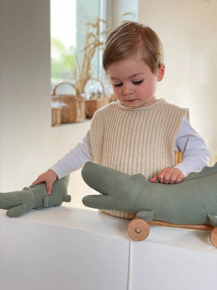 A young child in a cream knit vest and white shirt plays indoors with the Egmont Toys Pull-Along Croc, a green crocodile toy on wooden wheels. Soft natural light fills the room.
