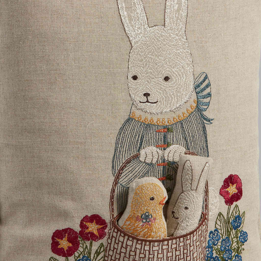 The Easter Bunny Pocket Pillow features embroidery of the bunny in a blue outfit with a basket containing a yellow chick and rabbit. Red flowers with green leaves border the bottom, making it ideal for festive décor.