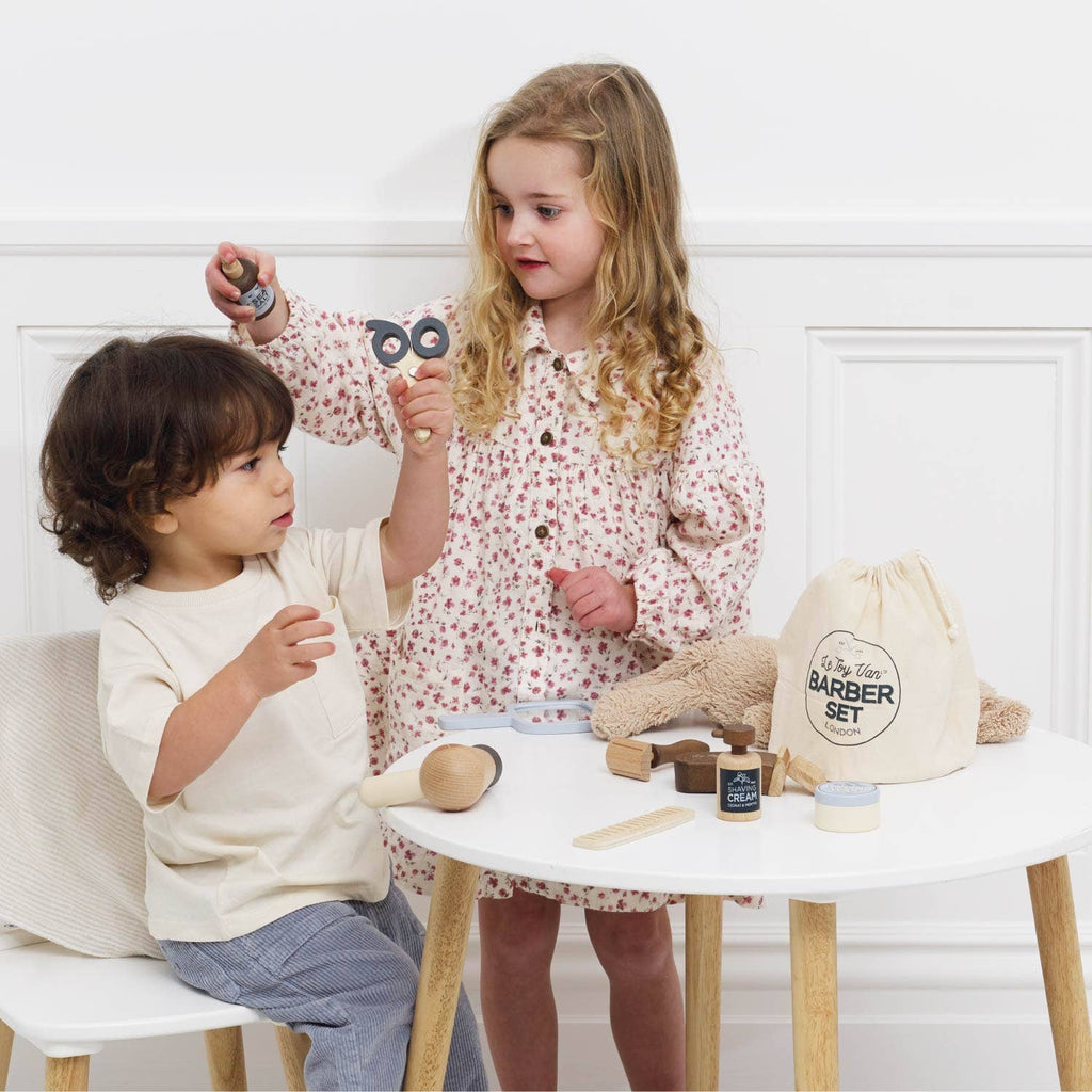 Two young children play at a small white table with the Wooden Hairdresser & Barber Kit, featuring a toy razor, brush, and barber bag. Dressed in bright colors, they pretend to cut hair with this charming plastic-free set (ships in one week).