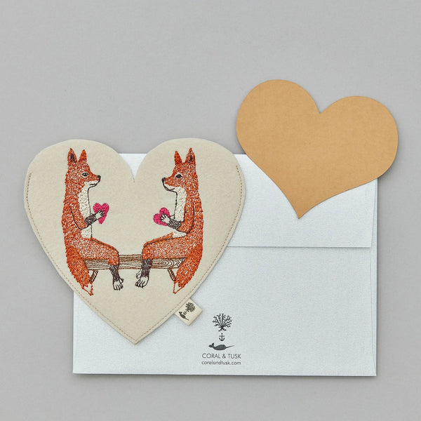 An embroidered Coral & Tusk Smitten Foxes felt envelope featuring two foxes holding pink flowers, with a smaller paper heart-shaped insert and an envelope. The card is on a light grey background.