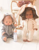 Two dolls dressed in hooded onesies with bunny ears sit on a white floor. The doll from the Tiny Harlow Bottled Milk and Juice Set wears gray and holds a magic bottle, while the other wears a peach outfit with curly hair. A chair is partially visible in the background.