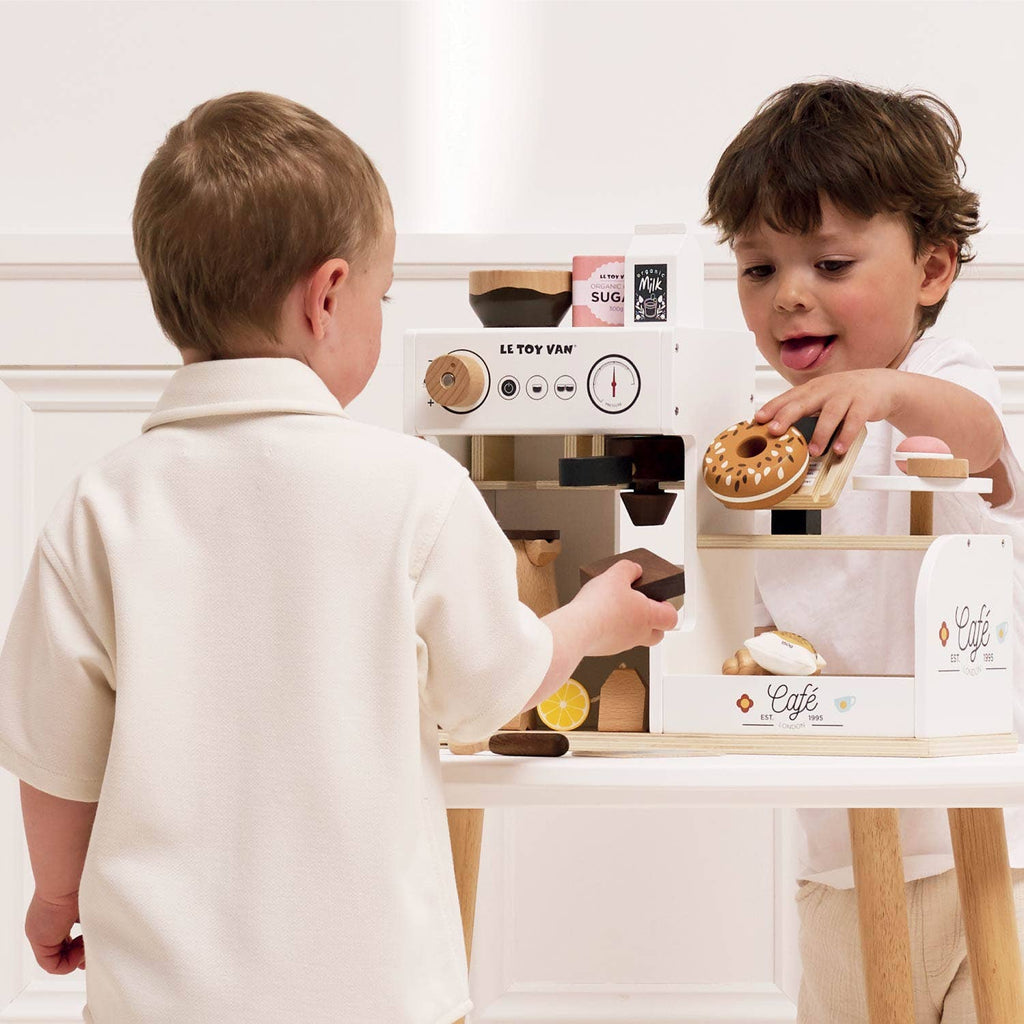 Two young children play with the Barista Café Shop—pretending to serve pastries and drinks. One holds a toy doughnut while the other uses the wooden coffee machine. Both wear light-colored clothes. Ships in one week.