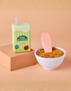 The Tiny Tummies Set - Alphabet Soup Bowl and Juice Box For Dolls, featuring a toy apple juice box and a bowl of toy alphabet soup with a large pink spoon, is displayed on a peach-colored surface, with the juice box positioned atop a cube-shaped platform.