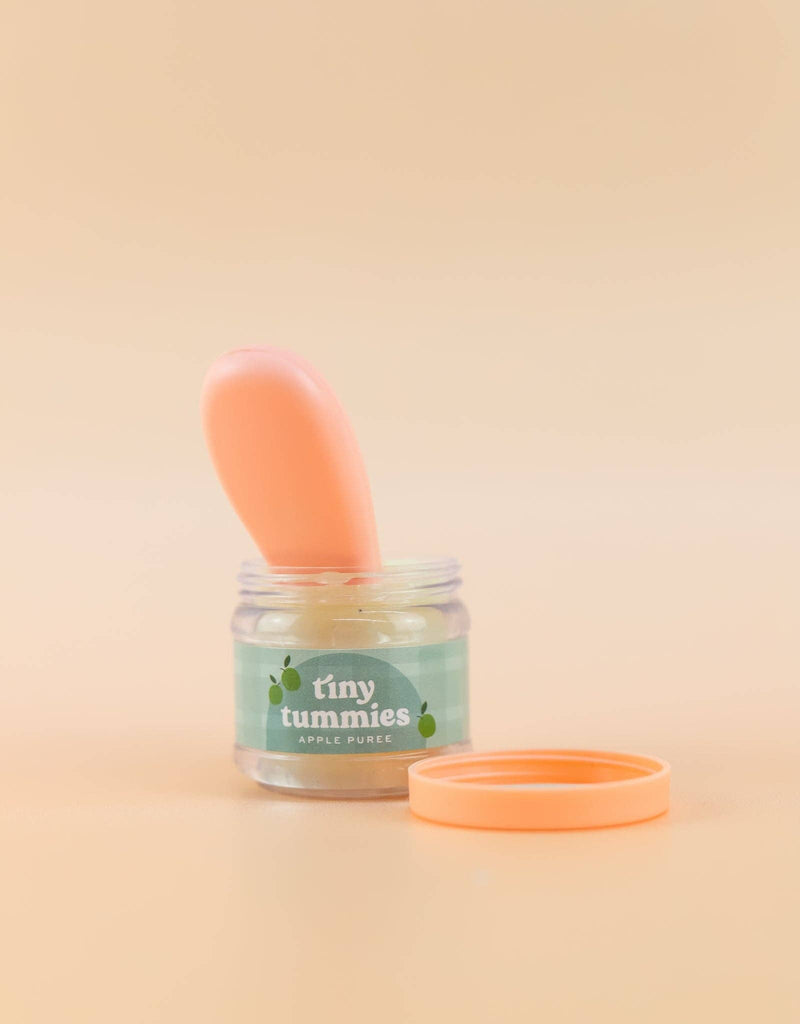 A small jar marked "Tiny Tummies - Apple jelly food" from Tiny Harlow, featuring a magic spoon within, sits on a neutral background. A light orange lid rests beside it, ready for your little doll's mealtime.