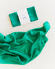 A packaged “Sarah's Silks Solid Color Playsilk - Emerald” rests above a flowing green silk fabric on a light background, ideal for open-ended play.