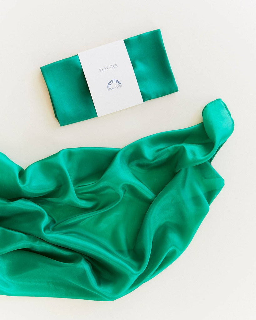 A packaged “Sarah's Silks Solid Color Playsilk - Emerald” rests above a flowing green silk fabric on a light background, ideal for open-ended play.