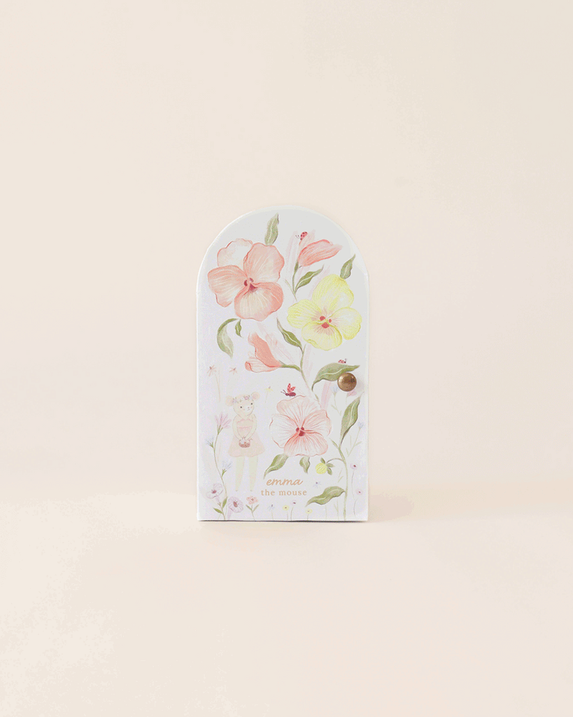 A small, arched white door featuring large pastel flowers, a brass knob, and an illustration of Tiny Emma the Mouse with "emma the mouse" text, handmade in Peru. Set against a plain beige backdrop. Product: Cuddle + Kind Tiny Emma the Mouse.