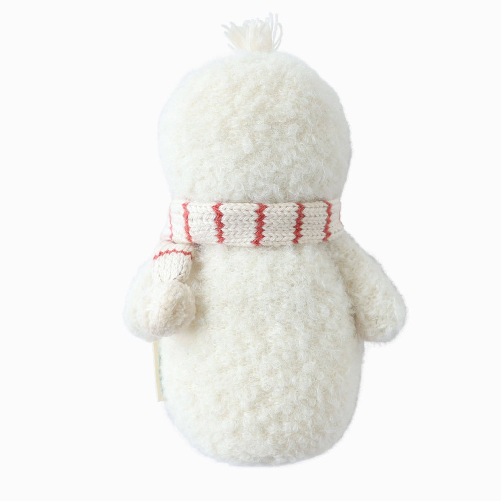 The Cuddle + Kind Baby Snowman - Candy Cane is a fluffy white plush toy with a round head, seen from behind, wearing a hand-knit red and white Peruvian alpaca yarn scarf—perfect as a winter collectible doll.