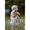 A young child in a light cap and shirt stands in a wildflower field, hugging the Senger Naturwelt Stuffed Animal - Small Sheep tightly, with blurred greenery in the background.