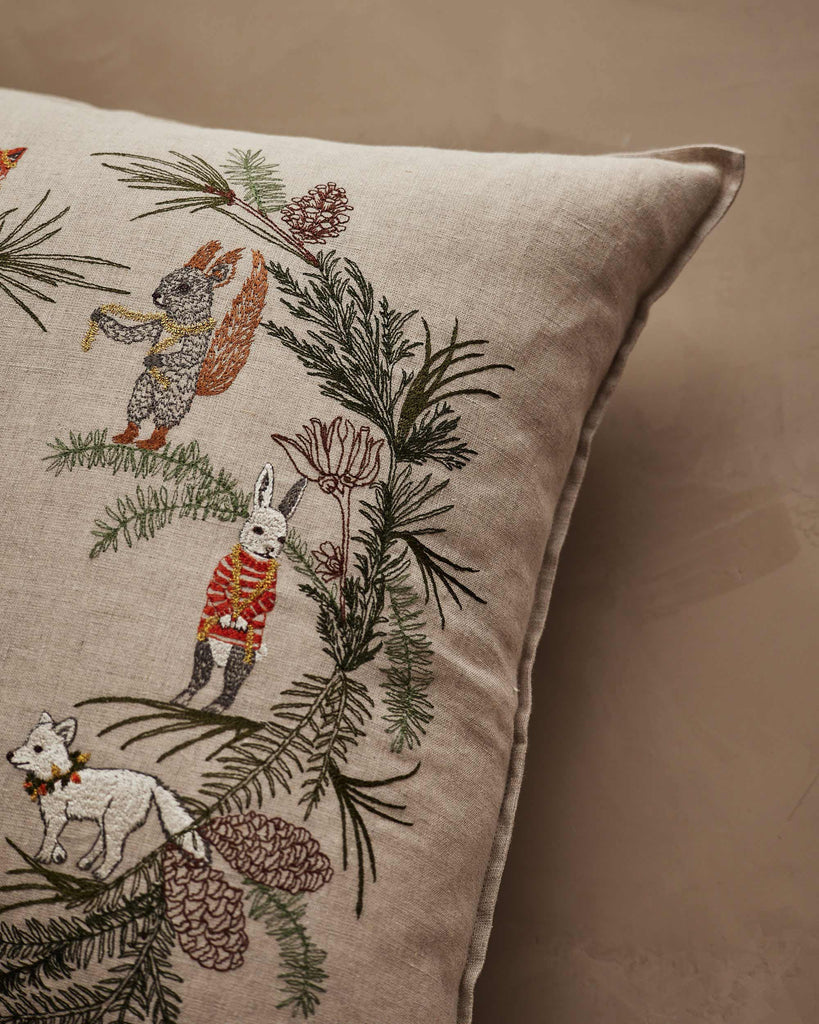 The Coral & Tusk Tree Trimmers Wreath Pillow features embroidered woodland animals—a squirrel, rabbit, and fox—adorned with pine branches and cones on a neutral beige background.