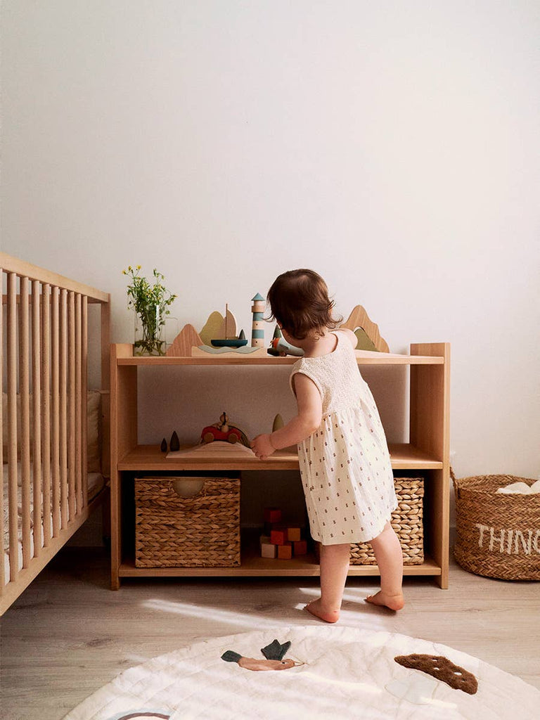 A young child in a cream dress stands in a cozy, minimalist nursery, reaching for the Sabo Concept Car with Lion on the shelf, surrounded by natural wood accents that inspire imaginative play.