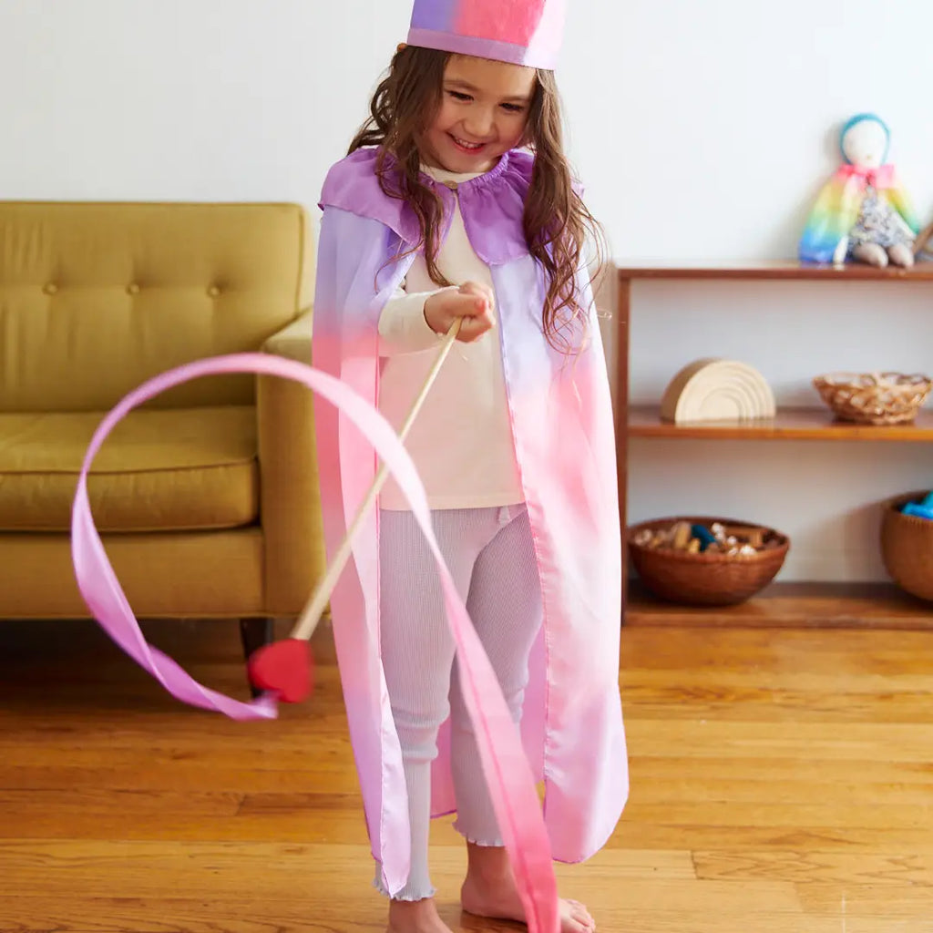 A young girl in a pink and purple outfit twirls Sarah's Silk Large Heart Streamer Wands in a living room, smiling happily while wearing a birthday hat.