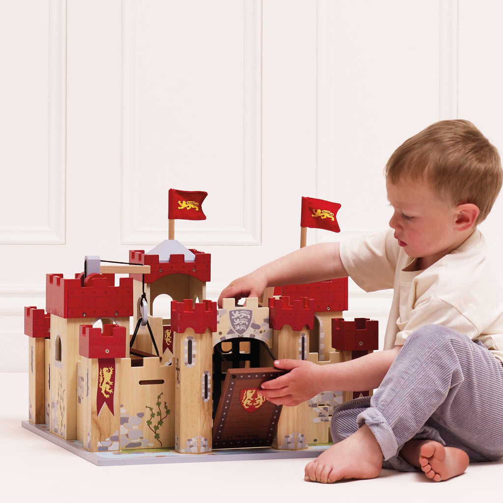 A young child sits on the floor, engaging in imaginative play with the Lionheart Wooden Castle, crafted from sustainable wood. Its bright red battlements and flags stand out against the natural finish as the child focuses intently, touching the castle’s drawbridge.