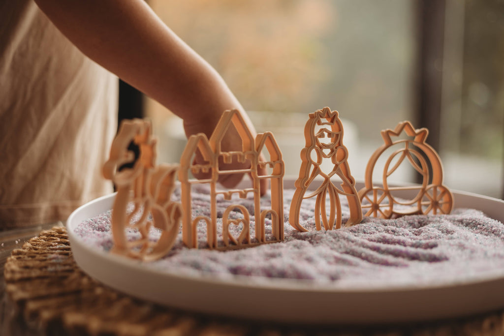 A child's hand reaches toward the Princess Cutter Set - Eco Playdough Tool—wooden, princess-themed figurines like a dragon, castle, princess, and carriage—displayed on a tray of purple sand and accompanied by eco-friendly playdough cutters and tools for imaginative storytelling.