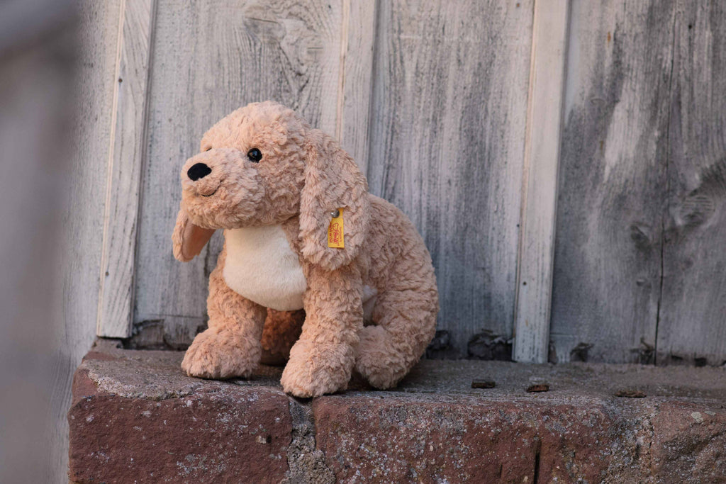 The Steiff Berno Goldendoodle Dog Plush, a brown toy with floppy ears, rests on a worn brick surface against a rustic wooden wall. It boasts the iconic Steiff button-in-ear tag and is fully machine washable, enhancing its charming appeal.