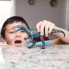 A young boy excitedly plays with a Schleich Baryonyx Realistic Dinosaur Toy with Movable Jaw on a granite countertop, his mouth open in delight, indoors with bright natural light streaming in the background.