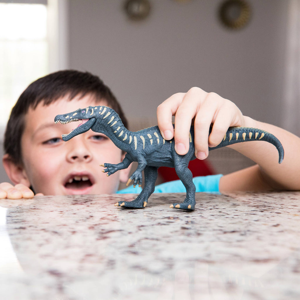 A young boy excitedly plays with a Schleich Baryonyx Realistic Dinosaur Toy with Movable Jaw on a granite countertop, his mouth open in delight, indoors with bright natural light streaming in the background.