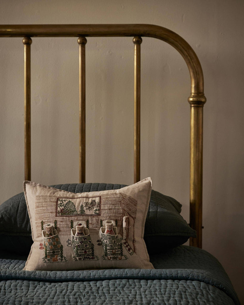 A brass bed frame with blue quilted bedding and pillows features the Coral & Tusk Three Little Bears Pocket Pillow, embroidered with three bears and a cottage scene, set against a plain beige wall. Ships in one week.