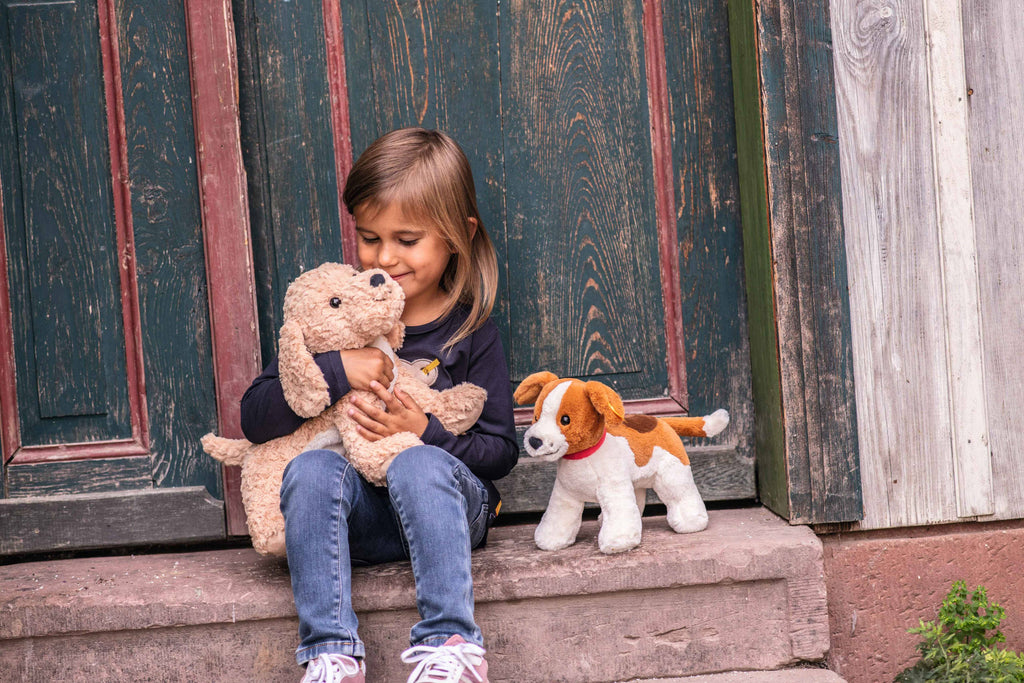 A young girl sits on a step in front of a wooden door, hugging a Steiff Berno Goldendoodle Dog Plush. Another stuffed animal is next to her. She smiles, wearing a black long-sleeve shirt and jeans.