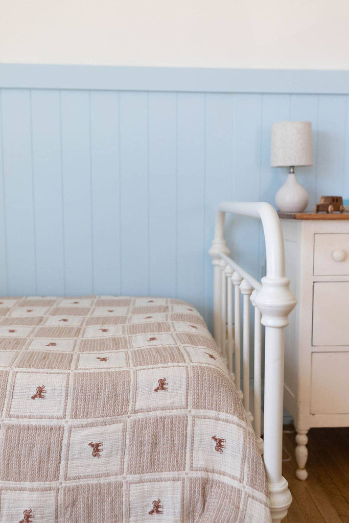 A cozy bedroom features a white metal bedframe, a white nightstand, a lamp, a toy car, and the Organic Cotton Twin Size Blanket - Pony with its beige and white checkered design and embroidered pony, set against a light blue paneled wall.