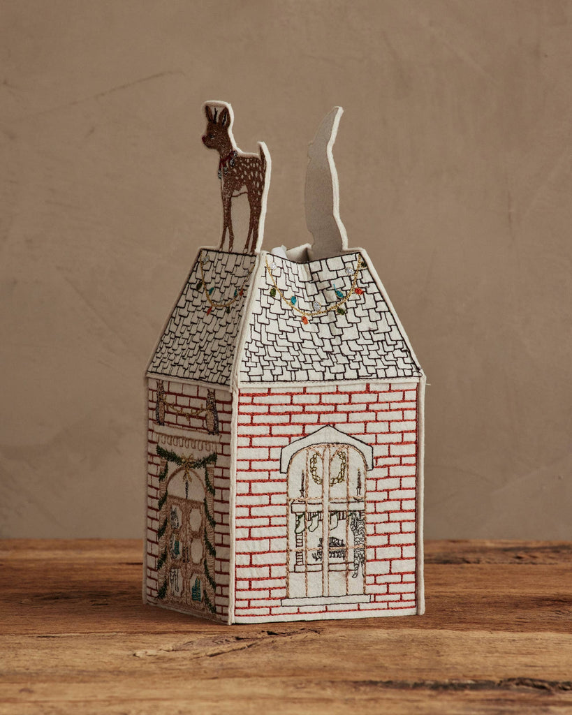 The Coral & Tusk Home for the Holidays Tissue Box Cover features a charming red brick house with embroidered windows, holiday lights, and a reindeer on a snowy roof—an ideal festive accent for your home.