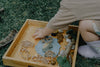 A child reaches into a wooden tray on grass, filled with playdough, stones, sticks, leaves, eco bubble wands, and the Pond Life Cutter Set – an eco-friendly playdough tool crafted from biodegradable PLA.