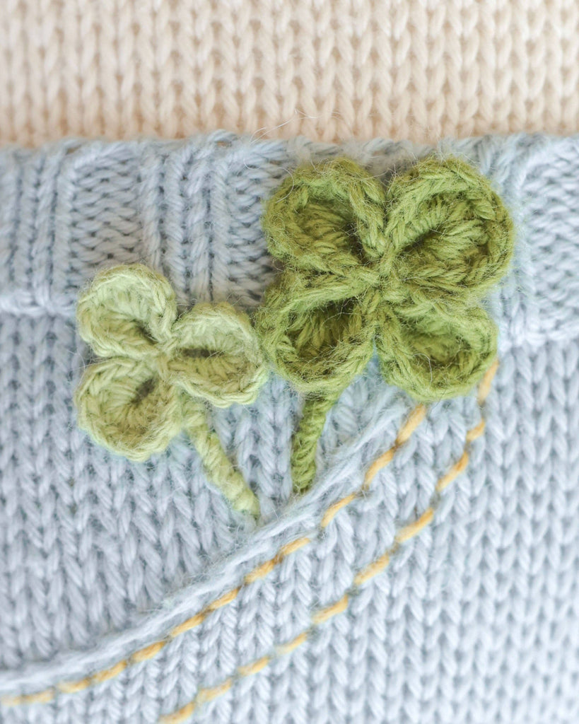 Close-up of light blue knitted fabric on the Cuddle + Kind Liam the Pony, handmade in Peru, featuring two green yarn-embroidered clovers and yellow stitched lines, set against a cream background.