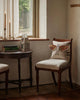 A person with butterfly wings sits by a window on a wooden chair with a beige cushion. Next to them, books, candles, and the Coral & Tusk Bat Wing Surprise Pillow create a cozy vintage vibe as warm sunlight streams in.