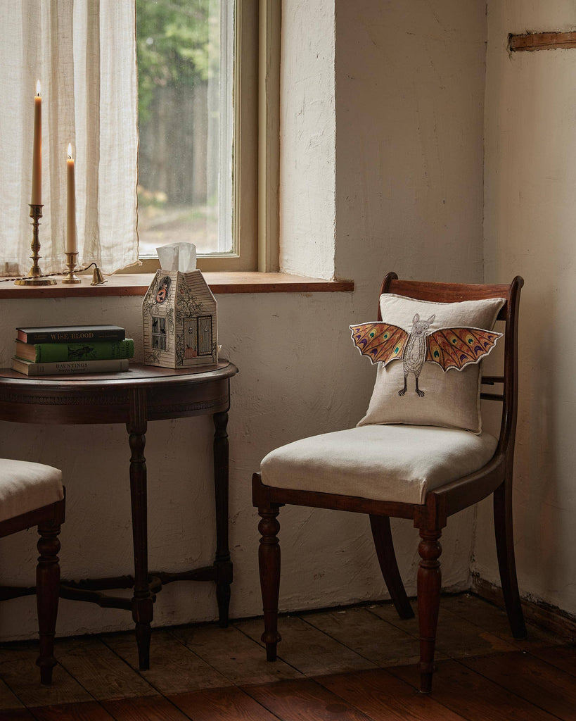 A cozy room with two wooden chairs, one featuring a pillow of a human figure with butterfly wings. A small table by the window holds candles, books, and a Coral & Tusk Haunted House Tissue Box Cover.