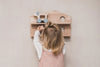 A young child with blonde hair is seen from behind as they interact with a handmade wooden wall-mounted shelf shaped like a house. The child, working on their fine motor skills, is reaching for a Wooden Puppy Push Toy placed on the top shelf. The background is a neutral-colored wall.