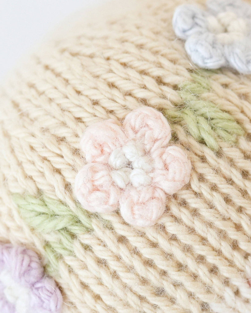 Close-up of Cuddle + Kind Tiny Emma the Mouse: a cream-colored, handknit fabric made in Peru, features embroidered light pink and lavender floral designs with green leaves.